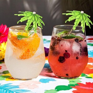 May include: Two stemless glasses filled with cocktails. One has an orange slice, the other berries. Both are garnished with green paper umbrellas and mint leaves. The drinks sit on a colorful floral patterned tablecloth.