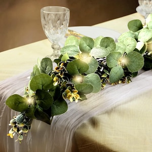 May include: A decorative eucalyptus garland with large, round green leaves and small yellow-green buds. The garland is illuminated with small, warm white lights, creating a soft glow. It is draped over a white tablecloth.