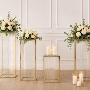 May include: Four decorative glass and gold stands of varying heights. The stands are rectangular with vertical ribbed glass panels and gold frames. Two stands hold bouquets of white roses and greenery. Candles are placed on the stands and in glass holders.