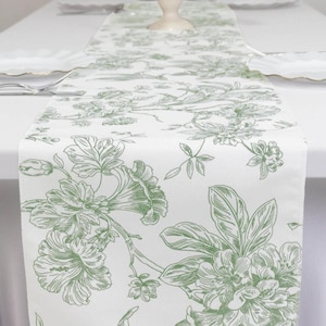 May include: White table runner with a green floral toile pattern. The runner features detailed botanical illustrations of flowers and foliage. The design is printed on a white background, creating a classic and elegant look. Perfect for adding a touch of sophistication to any dining table.