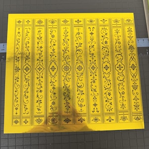 Puede incluir: Una hoja cuadrada de papel de aluminio dorado reflectante con un patrón repetitivo de diez columnas verticales. Cada columna contiene intrincados diseños de líneas negras de enredaderas florales, hojas en espiral y medallones geométricos.