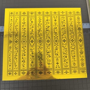 Puede incluir: Una hoja cuadrada de papel de aluminio dorado reflectante con un patrón repetitivo de diez columnas verticales. Cada columna contiene intrincados diseños de líneas negras de enredaderas florales, hojas en espiral y medallones geométricos.