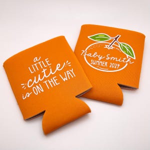 May include: Two orange foam can coolers. One features the white text 'A LITTLE CUTIE IS ON THE WAY'. The other displays a white graphic of an orange with leaves and the text 'Baby Smith SUMMER 2027' in white script and block lettering.