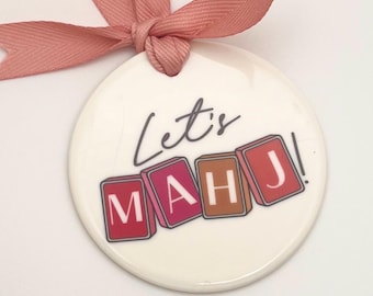 May include: A round, off-white ornament with the words "Let's MAH J!" printed on it. The letters MAH J are in colorful tile designs. A pink ribbon is attached at the top.