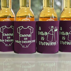 May include: Four purple beverage insulators with white text. Two insulators say "Cheers to Baby Davincent!" with a baby onesie graphic. Two insulators say "A Baby is Brewing!" with a bottle and a baby bottle graphic.
