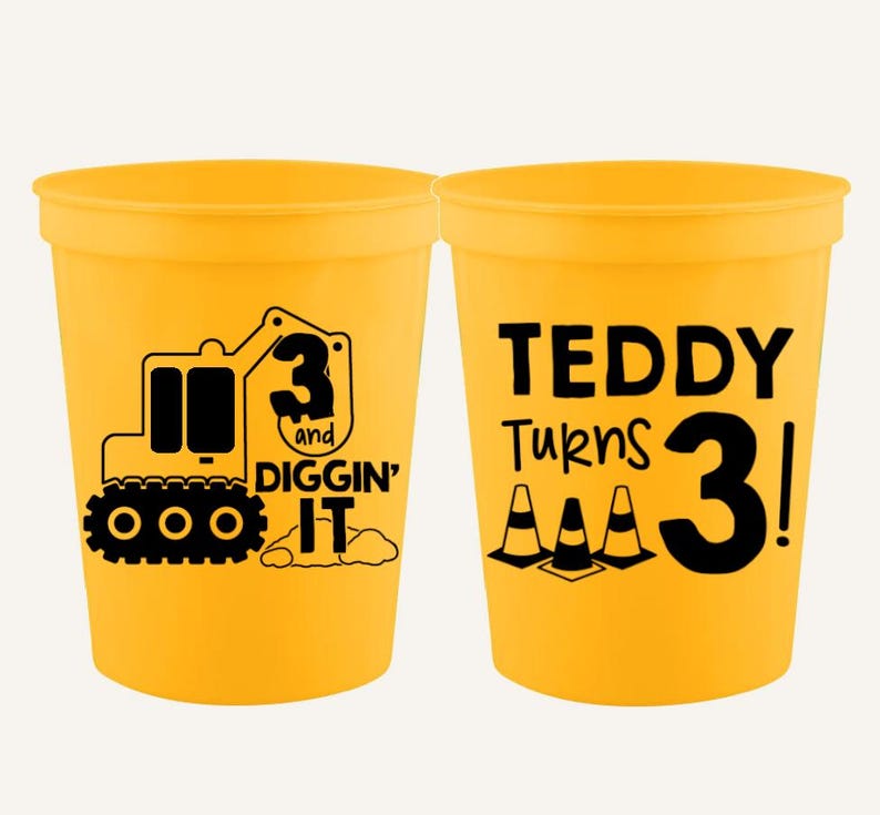 Op de afbeelding: Twee gele plastic bekers met zwarte grafische ontwerpen. E&eacute;n beker toont een graafmachine met de tekst 3 AND DIGGIN' IT. De andere beker bevat de tekst TEDDY TURNS 3! vergezeld van drie zwarte iconen van verkeerskegels in zwarte inkt.