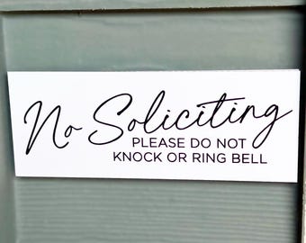Op de afbeelding: Een rechthoekig wit bord met zwarte tekst die No Soliciting leest in een cursief lettertype, gevolgd door de tekst PLEASE DO NOT KNOCK OR RING BELL in een strak, schreefloos hoofdlettertype, gecentreerd op het witte oppervlak.