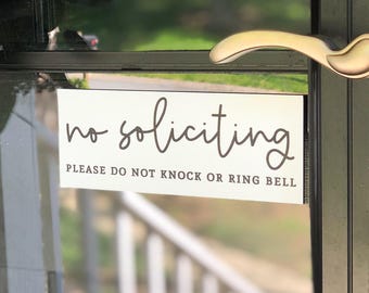 May include: A rectangular white sign with black text attached to a glass door. The sign features the words no soliciting in a cursive script font, followed by the text PLEASE DO NOT KNOCK OR RING BELL in a simple sans-serif capital font.