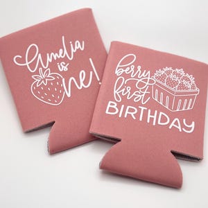 May include: Two dusty rose foam can coolers featuring white screen-printed text and graphics. One cooler displays the text Amelia is one! with a strawberry icon. The other cooler features the text berry first BIRTHDAY with a strawberry basket.