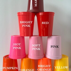 May include: A set of nine plastic cups in various shades of red, pink, orange, and yellow. The cups are stacked in a pyramid shape, with the top cup labeled "MAROON", the next row labeled "BRIGHT PINK" and "RED", the next row labeled "HOT PINK", "SOFT PINK", and "PINK", and the bottom row labeled "PUMPKIN", "ORANGE", "BRIGHT ORANGE", and "YELLOW".