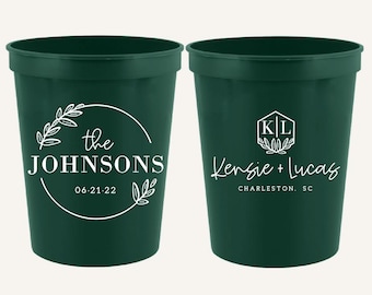 May include: Two dark green stadium cups featuring white printed text. The left cup displays "the JOHNSONS" with "06.21.22" inside a circular leaf wreath. The right cup shows "Kensie + Lucas" with "CHARLESTON, SC" and a hexagonal monogram "K|L".