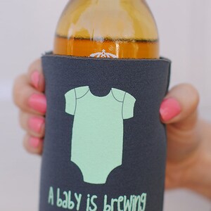 May include: A gray beverage insulator with a baby onesie graphic and the text "A baby is brewing".