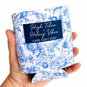 May include: A white foam can cooler featuring a blue floral pattern and a dark blue rectangular center panel. The text reads High Tides + Hilary Vibes CAPE COD | 2027 in white script and block lettering.