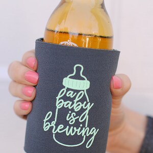 May include: A gray can cooler with a baby bottle graphic and the text "a baby is brewing" in green.