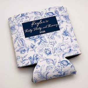May include: A white and blue floral print can cooler with the text "Kaylee's Thirty, Flirty, and Thriving 2026" on a navy blue rectangle. The cooler has a black interior and is designed to hold a beverage.