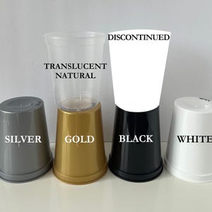 Op de afbeelding: Een verzameling plastic bekers in verschillende kleuren, waaronder zilver, goud, zwart en wit, samen met een doorzichtige natuurlijke beker. Tekstlabels identificeren de kleuren en geven aan dat de witte beker niet meer leverbaar is.