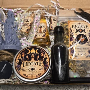 May include: A ritual kit box containing a purple Hecate statue, three small glass bottles with labels, a black liquid bottle, a round tin labeled Hecate Mini Ritual Kit, a black tea light candle, a bag of ritual blend herbs, a metal key, and a black moon charm.