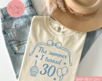 May include: Cream-colored t-shirt with the text "The summer I turned 30" in light blue, along with a cupcake, stars, and balloons. The shirt is paired with light-wash jeans, a necklace, and a tan hat. A sprig of dried flowers is also present.