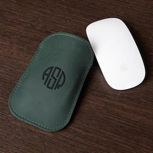 Puede incluir: Una funda de ratón de cuero verde oscuro con un monograma y un ratón inalámbrico blanco. La funda tiene un borde cosido y un monograma circular. El ratón es blanco con un logotipo de manzana plateada.