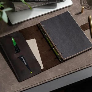 Peut inclure: Un classeur en cuir ouvert avec une couverture gris foncé et des anneaux argentés. Le classeur est ouvert, révélant un intérieur marron foncé avec une poche contenant une carte de crédit et une carte blanche. Un stylo et un crayon dorés sont à proximité.