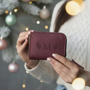 Könnte beinhalten: Eine weinrote Ledergeldbörse mit Reißverschluss. Die Geldbörse zeigt den Buchstaben "M" mit Blättermotiven. Die Geldbörse wird vor einem verschwommenen Hintergrund mit Weihnachtsdekorationen und Lichtern gehalten.