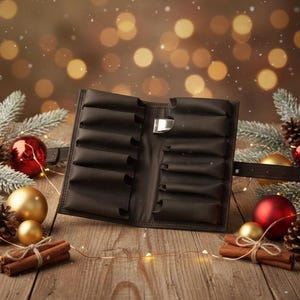 Puede incluir: Estuche de cuero negro abierto con múltiples compartimentos, asegurado por un cinturón. El estuche se muestra sobre una superficie de madera, rodeado de adornos navideños, piñas y palitos de canela. El fondo presenta luces bokeh borrosas.