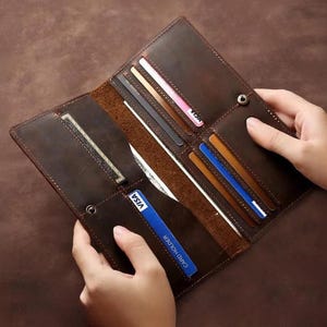 Könnte beinhalten: Offenes braunes Lederportemonnaie mit mehreren Kartenfächern und einem Druckknopfverschluss. Das Portemonnaie wird von zwei Händen gehalten, die den Inhalt zeigen.