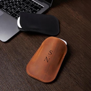 Op de afbeelding: Twee leren hoesjes voor een computermuis, één zwart en één bruin. De bruine hoes heeft de initialen "SN" gegraveerd. Een laptop is gedeeltelijk zichtbaar op de achtergrond, op een donker houten oppervlak.