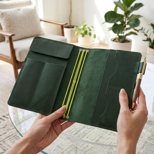 May include: A dark green leather notebook cover featuring three bright yellow elastic bands for securing inserts, multiple interior pockets for organization, and a wooden pen held in a side loop. The interior displays a smooth, matte finish.