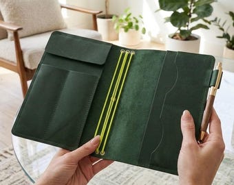 May include: A dark green leather notebook cover featuring three bright yellow elastic bands for securing inserts, multiple interior pockets for organisation, and a wooden pen held in a side loop. The interior displays a smooth, matte finish.
