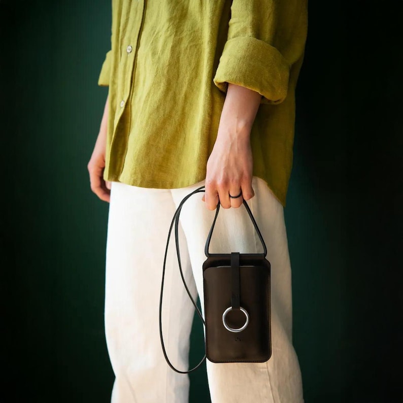 May include: A black leather crossbody phone bag with a silver ring detail. The bag has a long, thin strap and a flap closure. The person is wearing a green shirt and white pants.