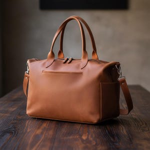 May include: A tan leather duffel bag featuring two top handles, a detachable shoulder strap with silver-toned metal hardware, a top zipper closure, and a horizontal zippered pocket on the front panel with a side pocket on the right side.