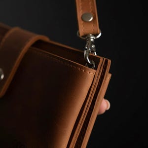 以下が含まれることがあります： 茶色の革製財布のクローズアップ。お揃いのストラップとシルバー色の留め金が付いています。財布には複数のコンパートメントとボタン留めが付いています。ストラップはシルバー色の留め金で財布に取り付けられています。