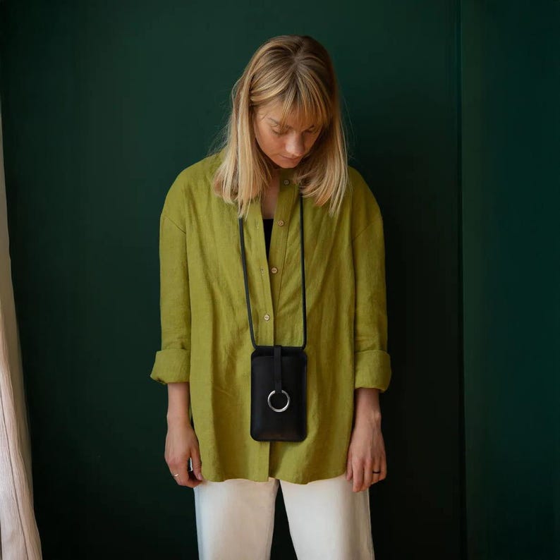 May include: A black leather phone case with a silver ring detail, worn as a crossbody bag. The case hangs from a black cord. The person is wearing a light green button-down shirt and white pants.