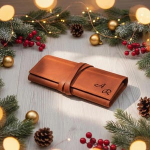 May include: A brown leather roll-up case with a strap closure, personalized with the initials "A R". The case is surrounded by Christmas decorations, including pine branches, red berries, gold ornaments, and pine cones.