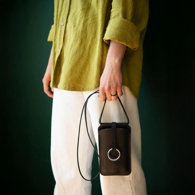 May include: A black leather phone pouch with a long thin shoulder strap and a silver metal ring closure on the front. The pouch is rectangular with rounded corners and a vertical strap that secures the top flap through the silver ring hardware.