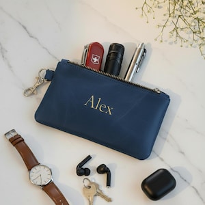 Pode incluir: Uma bolsa de couro azul marinho com o nome "Alex" em letras douradas, contendo um canivete suíço vermelho, uma lanterna preta e uma caneta prateada. Um relógio de pulso, fones de ouvido, chaves e um estojo de carregamento preto também são visíveis.