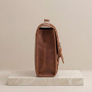 May include: A side view of a brown leather satchel bag featuring a structured rectangular body, a top handle with silver-toned metal hardware, and a front flap secured with a vertical strap and a silver-toned metal buckle closure mechanism.