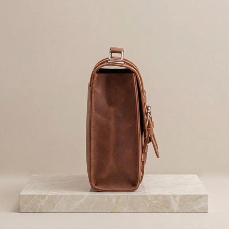 May include: A side view of a brown leather satchel bag featuring a structured rectangular body, a top handle with silver-toned metal hardware, and a front flap secured with a vertical strap and a silver-toned metal buckle closure mechanism.