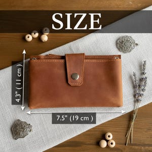 以下が含まれることがあります： スナップ留めの茶色の革製財布。長さ19cm、高さ11cm。財布の上には「SIZE」の文字が表示されています。財布は薄い灰色の表面に置かれています。