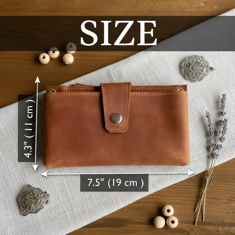 以下が含まれることがあります： スナップ留めの茶色の革製財布。長さ19cm、高さ11cm。財布の上には「SIZE」の文字が表示されています。財布は薄い灰色の表面に置かれています。