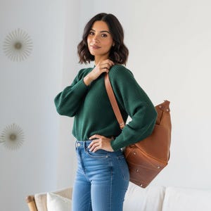May include: A dark green long-sleeved ribbed sweater paired with blue denim jeans and a tan leather shoulder bag featuring silver-toned metal rivets and a wide strap. The sweater has a crew neckline and a textured knit pattern throughout the fabric.