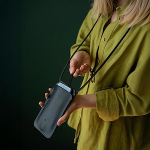 May include: A black leather phone case with a black strap. The phone case is rectangular and has a small, triangular logo on the front. The strap is thin and adjustable. The phone case is being held by a person wearing a green shirt.