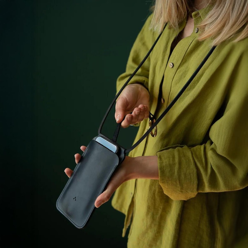 May include: A black leather phone case with a black strap. The phone case is rectangular and has a small, triangular logo on the front. The strap is thin and adjustable. The phone case is being held by a person wearing a green shirt.