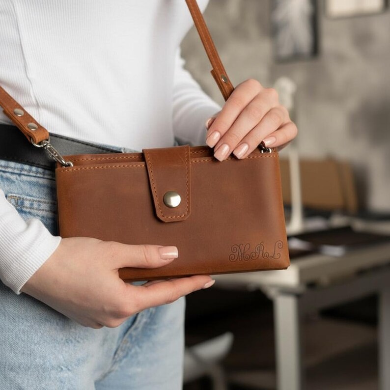 以下が含まれることがあります： フラップ開閉とシルバーのスナップが付いた、茶色の革製クロスボディバッグ。バッグには、長くて調節可能なストラップと、小さな刻印モノグラムが付いています。バッグは人が持っています。