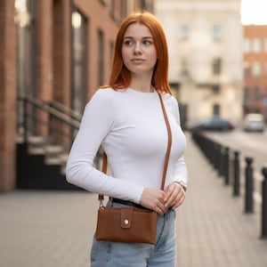 以下が含まれることがあります： 白い長袖シャツと薄い青色のジーンズを着た女性が、ボタン留めの茶色の革製クロスボディバッグを持っています。バッグには細い長いストラップが付いています。背景はレンガ造りの建物がある街並みです。