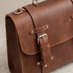 May include: A brown leather satchel featuring a top handle and a front flap secured with a vertical strap and a silver-toned metal buckle. The bag has visible stitching, smooth leather texture, and silver metal rivets securing the strap components.