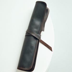 May include: A rolled-up, dark brown leather case with a matching strap wrapped around it. The case has a stitched border and is designed to hold items. The background is a solid white surface.