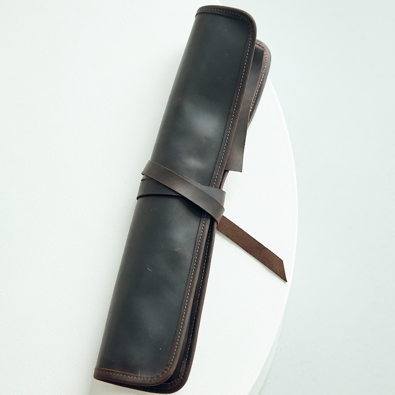May include: A rolled-up, dark brown leather case with a matching strap wrapped around it. The case has a stitched border and is designed to hold items. The background is a solid white surface.