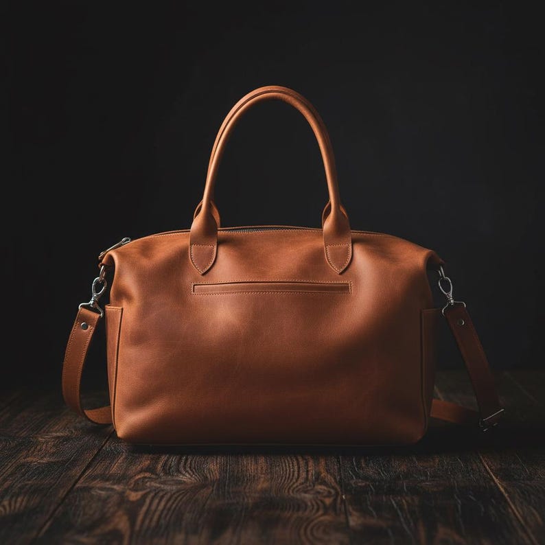 May include: A tan leather handbag featuring two top handles, a horizontal zippered pocket on the front, and detachable side straps with silver-toned metal hardware. The bag has a structured rectangular base and a smooth, matte finish throughout.